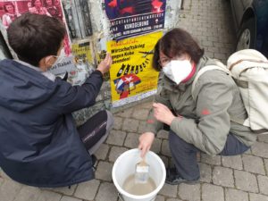 zwei Frauen kleben ein Plakat an eine Wand. Auf dem Plakat steht "Wirtschaftskrieg gegen Kuba beenden!"